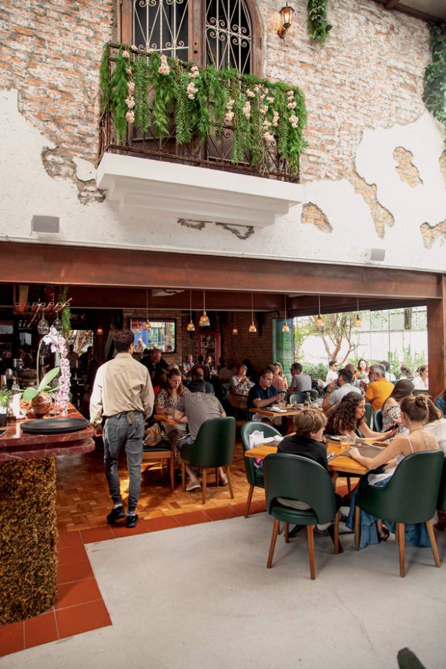Fachada e ambiente_6.jpg Entrada de restaurante com primeiro andar aberto, paredes brancas e floreira sobre a entrada, com clientes dentro e em mesas do lado de fora