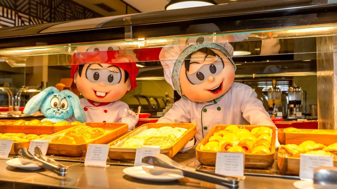 Pessoas vestidas como os personagens Mônica e Cebolinha com avental e touca de padeiro. Eles estão atrás de uma mesa cheia de cestas de pães para se servir