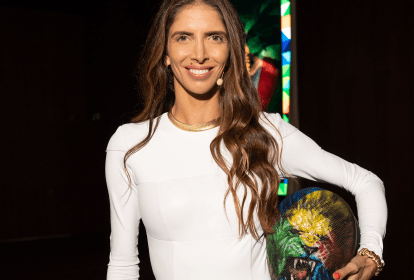 Nathalie Moellhausen posa com roupa branca justa no corpo e segura máscara debaixo do braço com desenho de leão colorido. Sorri e tem cabelos castanhos longos.