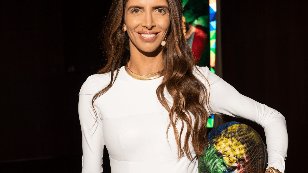 Nathalie Moellhausen posa com roupa branca justa no corpo e segura máscara debaixo do braço com desenho de leão colorido. Sorri e tem cabelos castanhos longos.