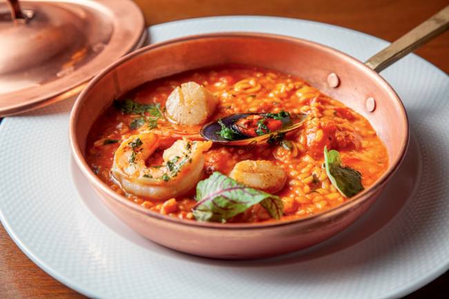 Risoto de frutos do mar Lvtetia Prato de porcelana com frigideira de bronze apoiado, contendo risoto de frutos do mar onde se veem camarões e mexilhões