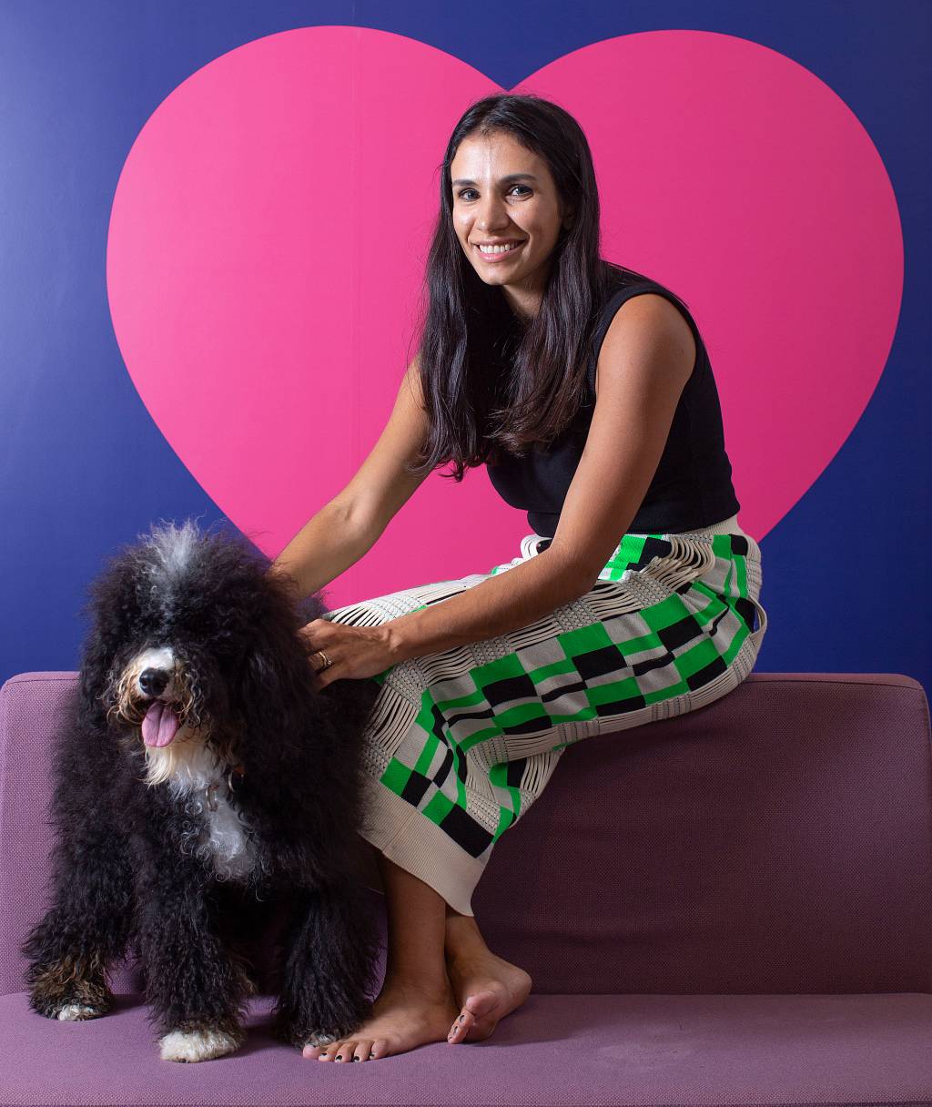 Talita Lacerda (Claudio Gatti-Divulgação) Talita Lacerda, CEO da Petlove, posa sentada com cachorro preto ao lado e fundo com coração rosa. Tem cabelos pretos longos e veste jeans clara.