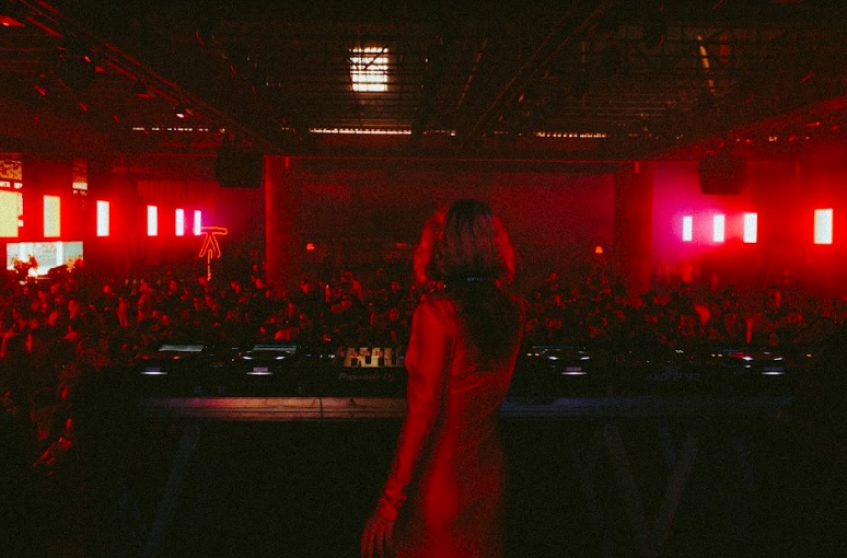 DJ de costas comanda som com pista lotada à frente. Foto tem tons em vermelho.