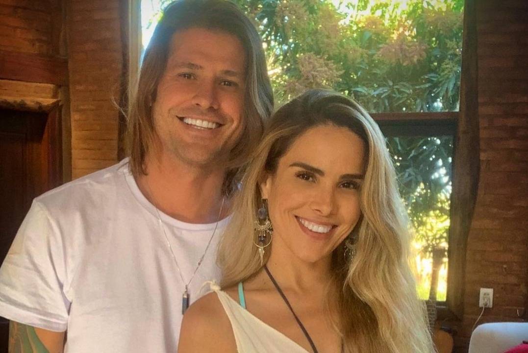 Foto de Dado Dolabella e Wanessa Camargo, posando, abraçados, olhando para a câmera e sorrindo. Ambos usam camisetas brancas