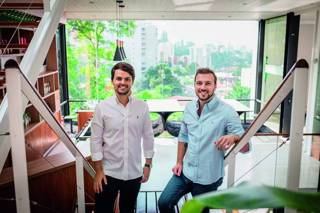 Nômade Festival Imagem mostra dois homens sorrindo,, um de camisa branca e o outro de camisa azul, em sala de apartamento