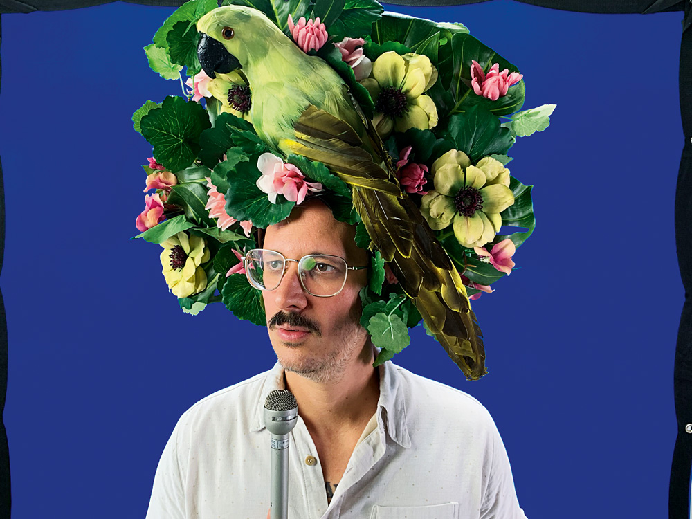 Imagem mostra homem de óculos e bigode com chapéu com flores e papagaio verde