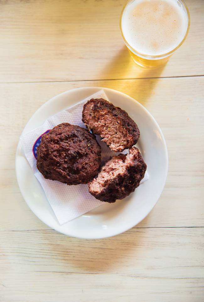 Dois bolinhos de carne, um partido ao meio, sobre guardanapo em prato branco, com um copo de chopp ao lado
