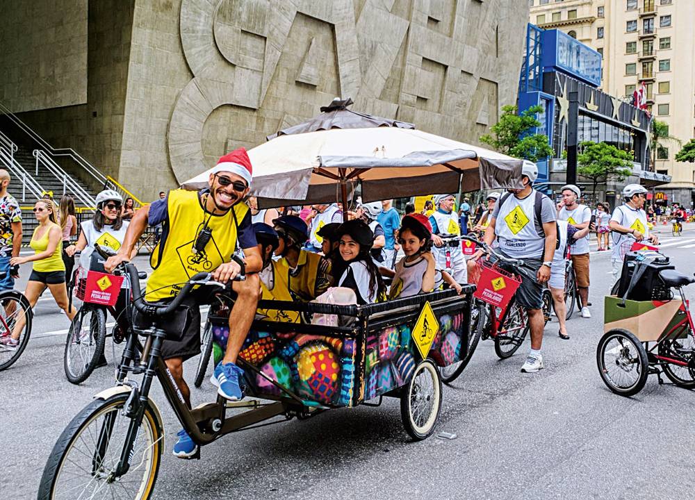 Pessoas pedalam na Avenida Paulista. No centro da foto, está um monitor com um pequeno trailer aberto que carrega crianças