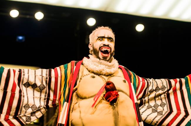 O ator Douglas Novais estende os braços e sorri no palco. Ele usa lentes de contato chamativas, dentes afiados e figurino bizarro e colorido