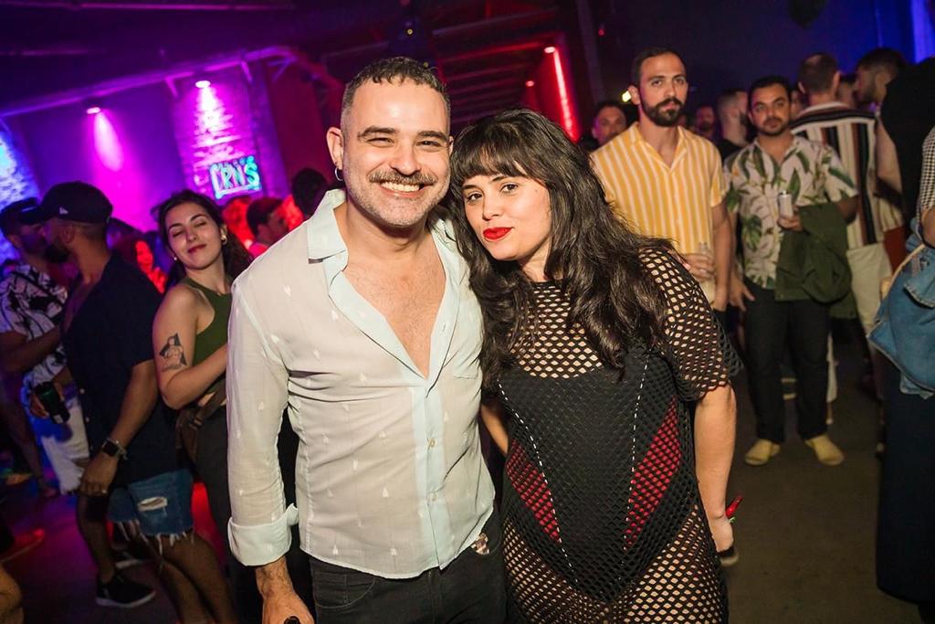 Dany Pita e Rodrigo Brandão posam lado a lado em festa. Ele veste camisa branca com botões abertos no peito e ela veste uma roupa preta bem ajustada ao corpo.