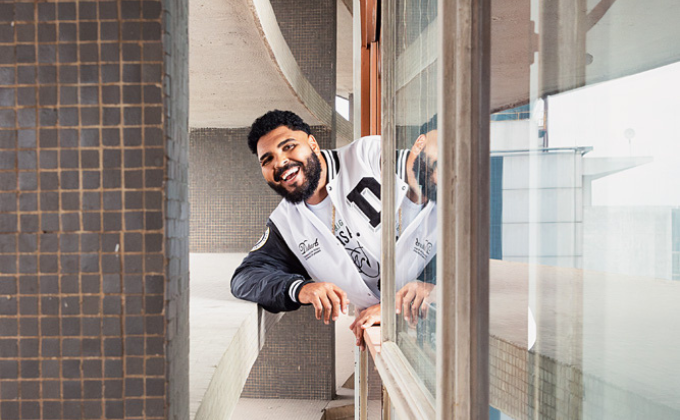 Imagem mostra homem sorrindo apoiado em janela, com o braço para fora