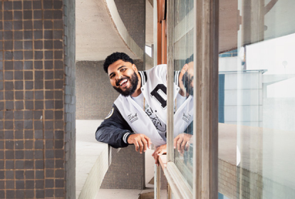 Imagem mostra homem sorrindo apoiado em janela, com o braço para fora