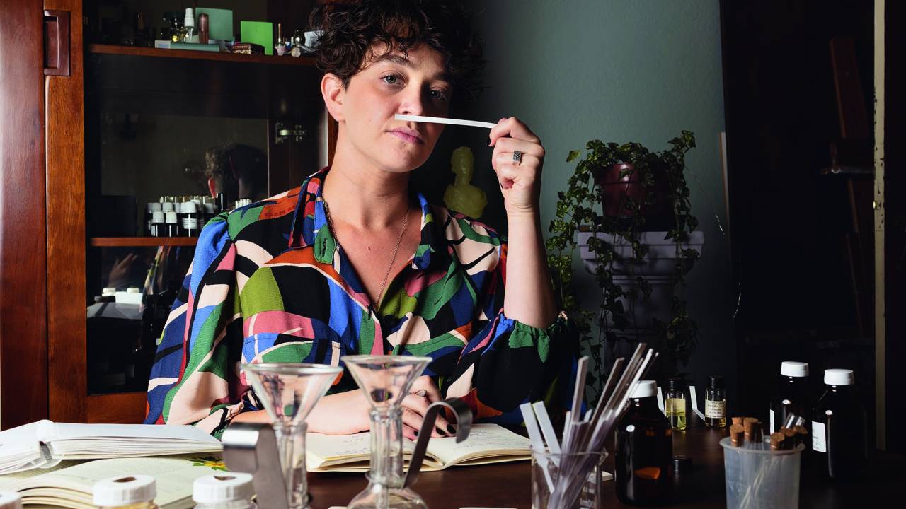 Mulher com cabelos curtos, branca, de camisa de manga curta estampada, segura uma fita olfativa próximo ao nariz, em frente a uma mesa de trabalho enquanto encara a câmera