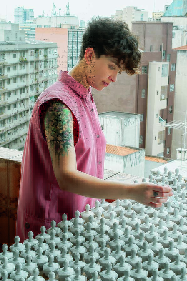 Karola Perfumação perfume concreto Mulher com camisa rosa sem mangas e cabelos curtos e tatuagem no ombro direito aplica essência em peças idênticas de concreto no formato de um frasco de perfume, dispostas sobre uma mesa. São muitos frascos, que preenchem a mesa toda.
