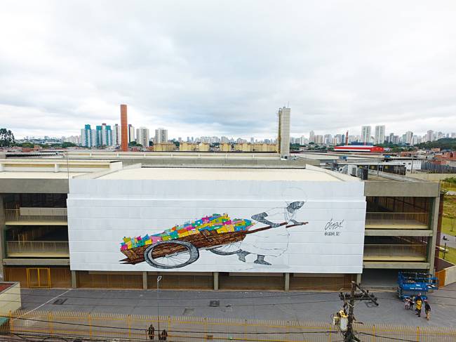 DCIM100MEDIADJI_0052.JPG Foto mostra grafite na empena de prédio com senhora vestida de branco, levando carrinho com pequenas casas