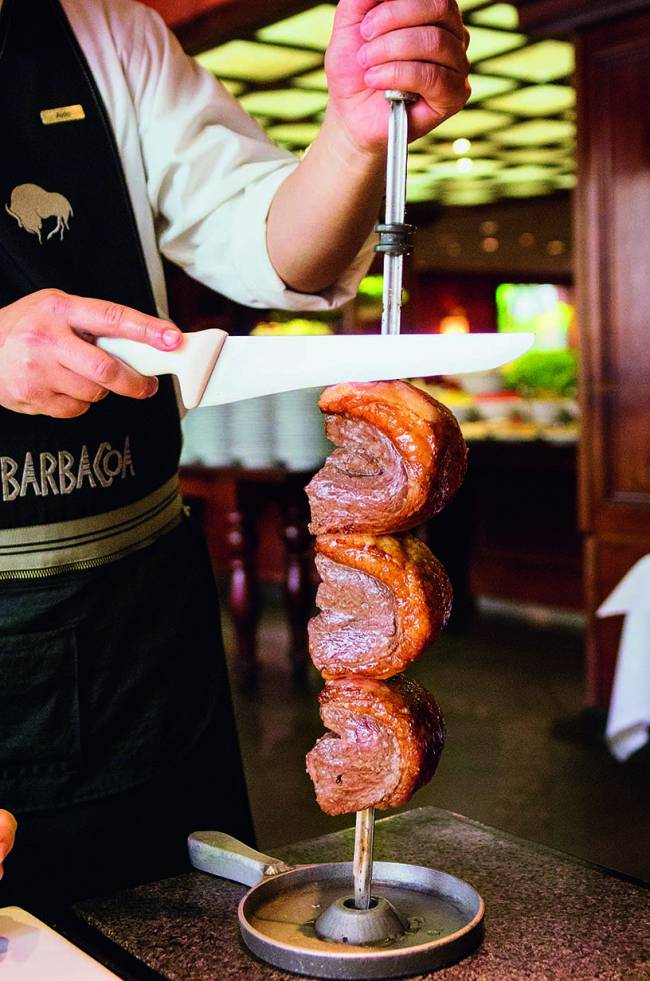 Picanha Barbacoa Itaim Pessoa vestida de garçom segurando espeto de picanha e faca par cortar fatia