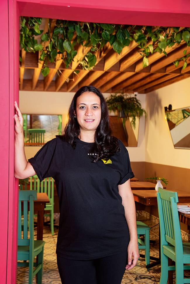 Mulher branca de cabelos pretos na altura dos ombros com camiseta preta de manga curta e apoiada num batente de porta com uma das mãos