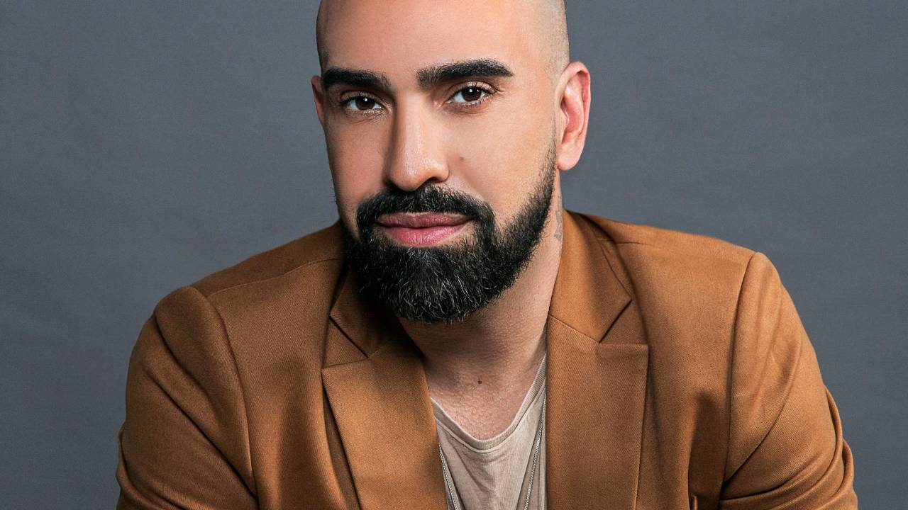 Homem branco e careca com barba preta encara a câmera. Veste blazer marrom.