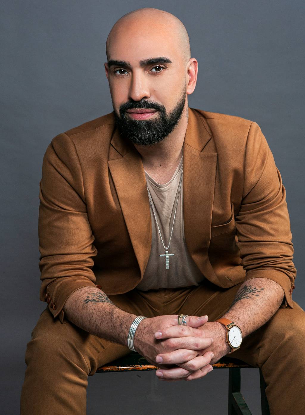 Homem branco e careca com barba preta encara a câmera. Veste blazer marrom.