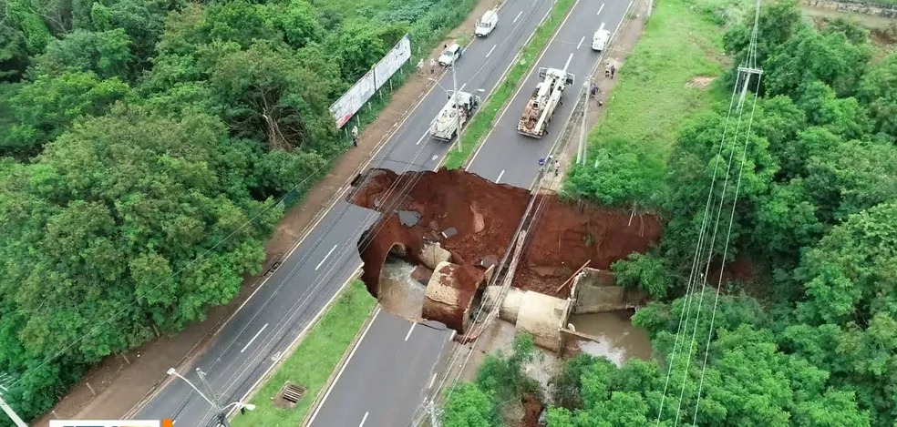 Foto de cratera ocupando quatro faixas em ambos os sentidos. Carro de bombeiros aparece parado em cima. Árvores em volta