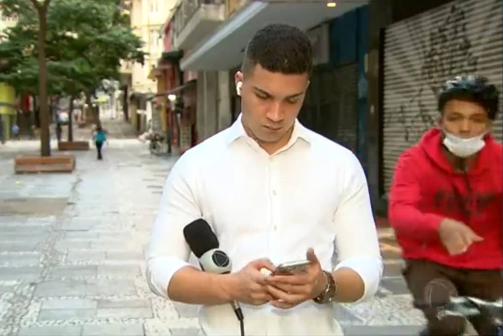 Imagem mostra homem de camisa branca, olhando o celular, e homem em bicicleta atrás