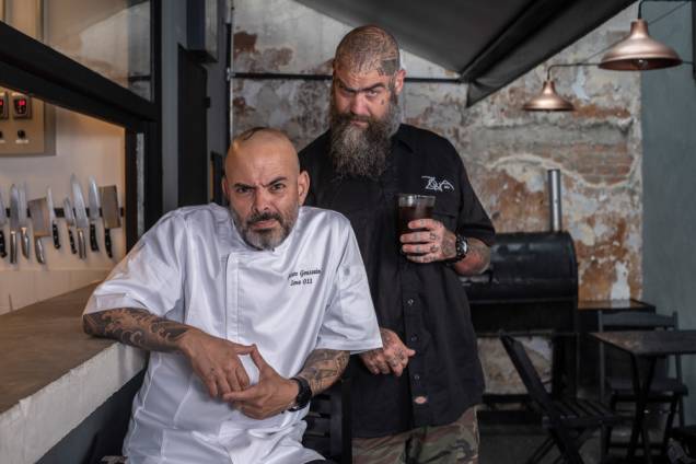Um homem careca de camiseta branca de mangas curtas e homem também careca de barba longa com camiseta preta, ambos tatuados, olham para a câmera de forma descontraída, fazendo careta Um homem careca de camiseta branca de mangas curtas e homem também careca de barba longa com camiseta preta, ambos tatuados, olham para a câmera de forma descontraída, fazendo careta