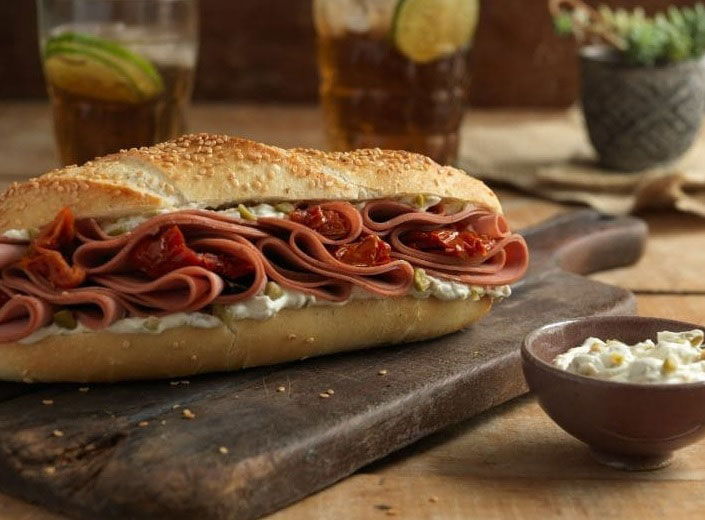 Sanduíche de mortadela no pão baguete com gergelim sobre tábua de madeira ao lado de cumbuca de madeira contendo um molho esbranquiçado