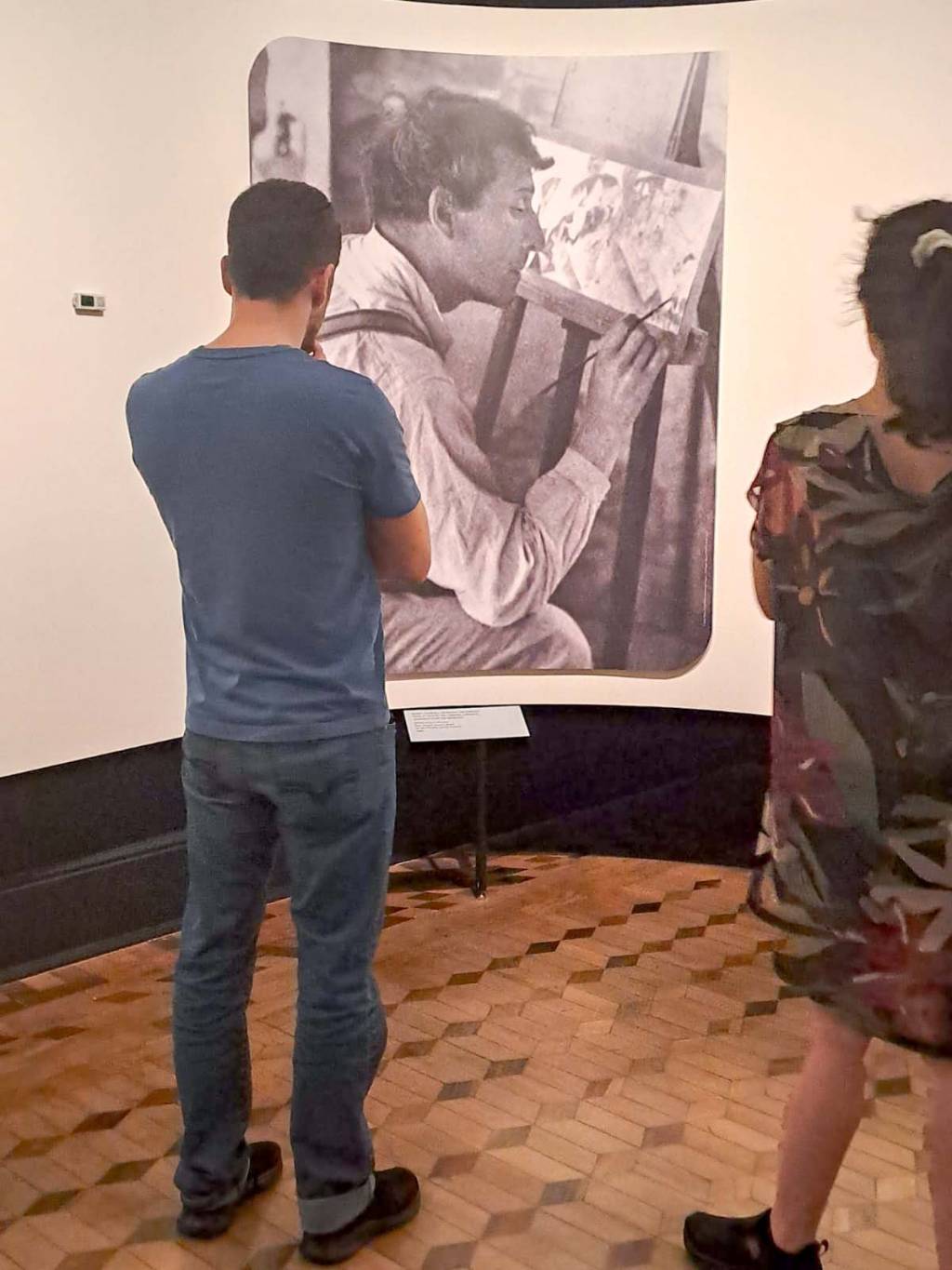 expo marc chagall bh Um homem e uma mulher olham para uma foto em preto e branco de Marc Chagall pintando. Eles estão de costas