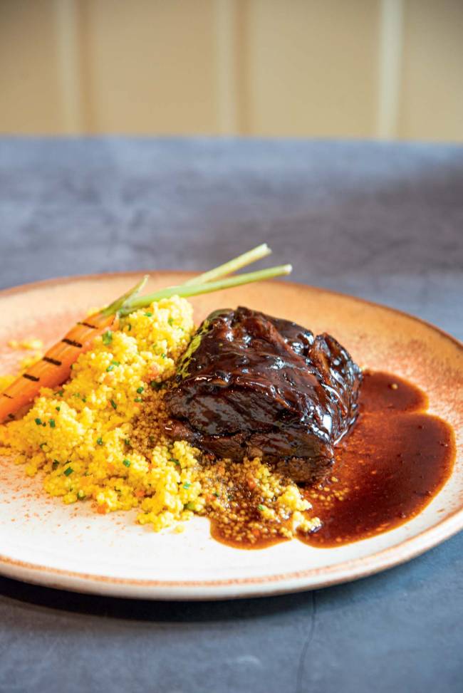 Prato de cerâmica sobre mesa azul, com paleta de cordeiro, cuscuz marroquino temperado e uma minicenoura com marcas de grelha