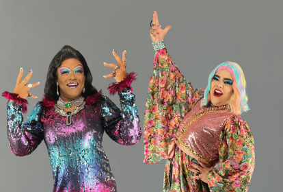 Duas drag queens estendem as mãos e posam sorridentes. A da esquerda é negra e veste vestido nas cores roxo, azul e rosa. A da direita é branca, com peruca branca e azulada, e veste peça florida.