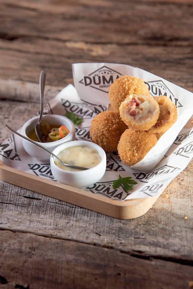 Bolinho de queijo com bacon Dümai Charcutaria bandeja de madeira com dois recipientes de molho, papel forrando com o logotipo da Dümai e bolinhos empilhados, um deles partido ao meio