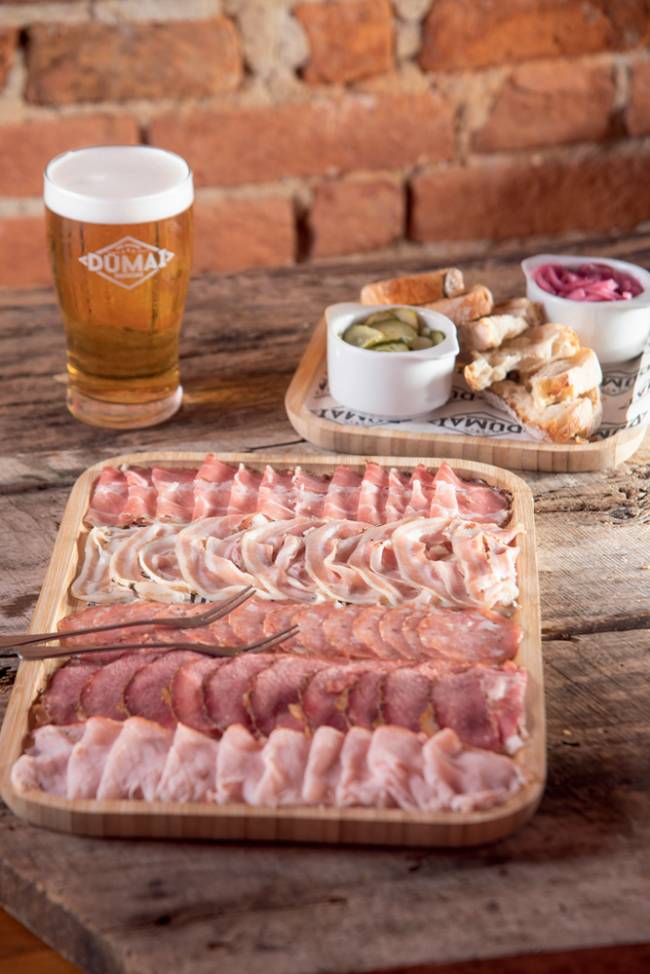 Tábua Mista Dümai Charcutaria tábua de madeira com diversas carnes curadas e defumadas, ao lado de tábua menor com dois tipos de picles e pão em fatias e copo de chopp