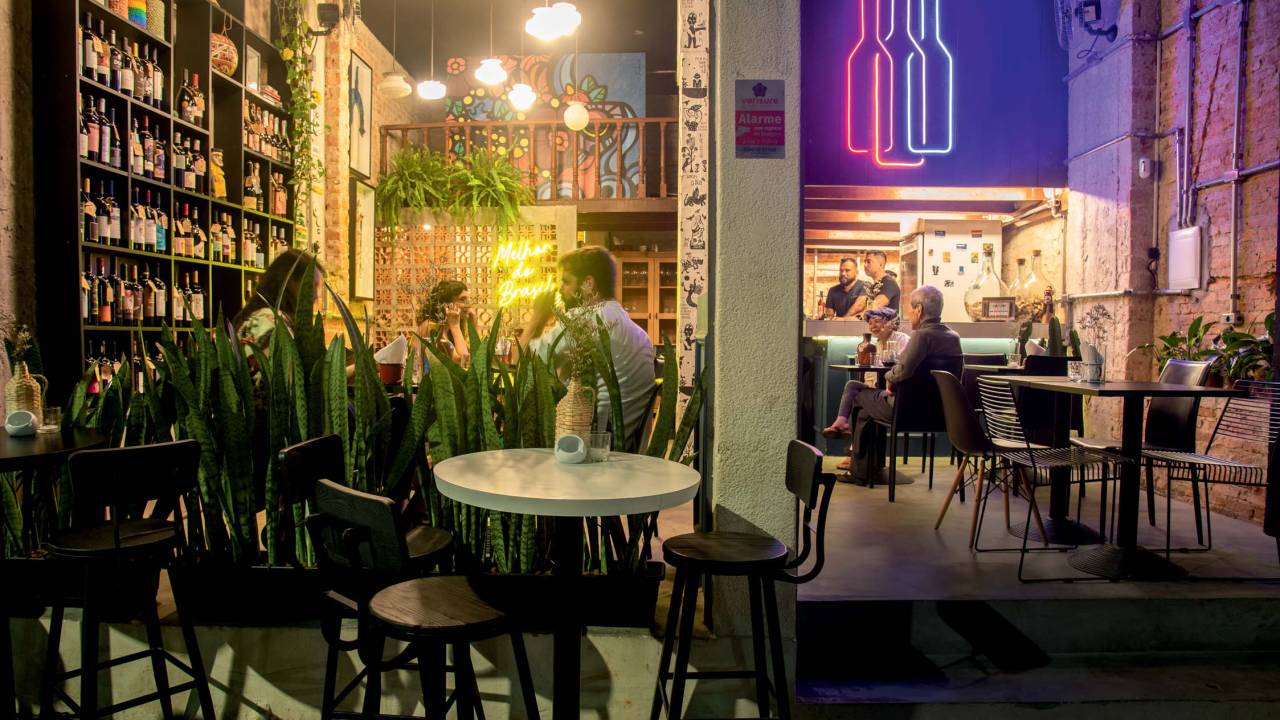 Fachada de espaço com luz amarela, tijolinhos à vista e mesinhas ao ar livre, com neons no formato de garrafas