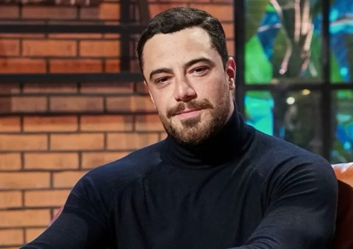 Foto de homem branco com barba e topete sorrindo, usando camiseta preta gola alta, em frente a parede de tijolinhos