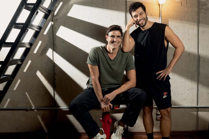 Reynaldo Gianecchini e Bruno Fagundes posam sorrindo para uma foto. Reynaldo está sentado em um banco vermelho, usa óculos, camiseta cinza e calça jeans. Bruno está em pé e apoia o cotovelo no ombro dele. Ele usa uma regata preta e um short preto. Ambos são homens brancos, de cabelos e barba escuros