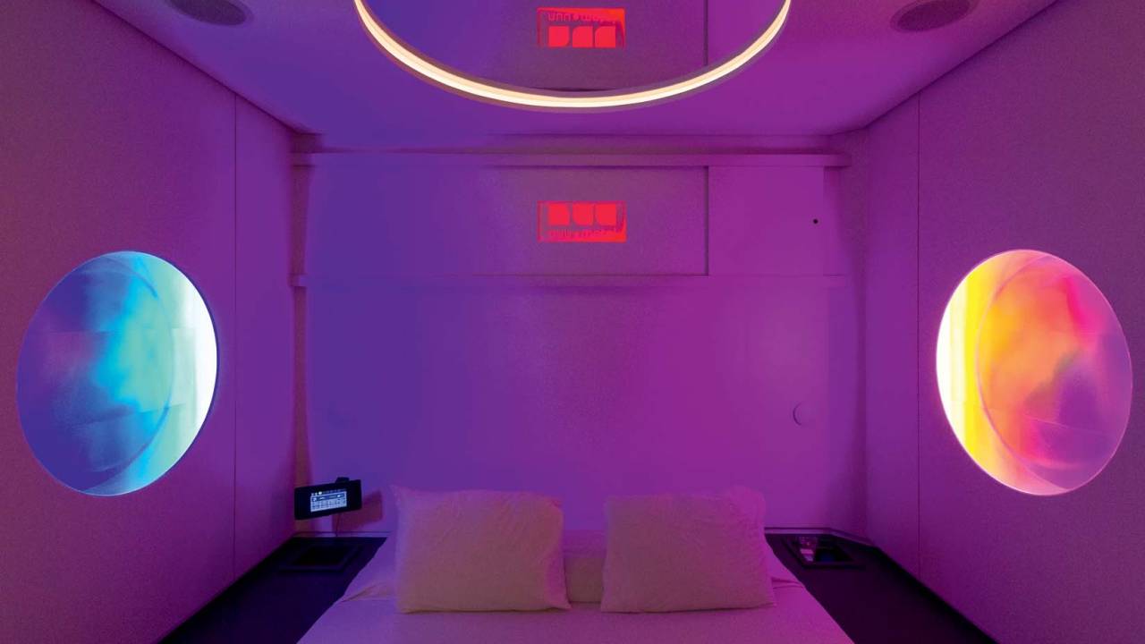 Foto mostra cama em quarto de motel com luz LED em tom roxo pastel