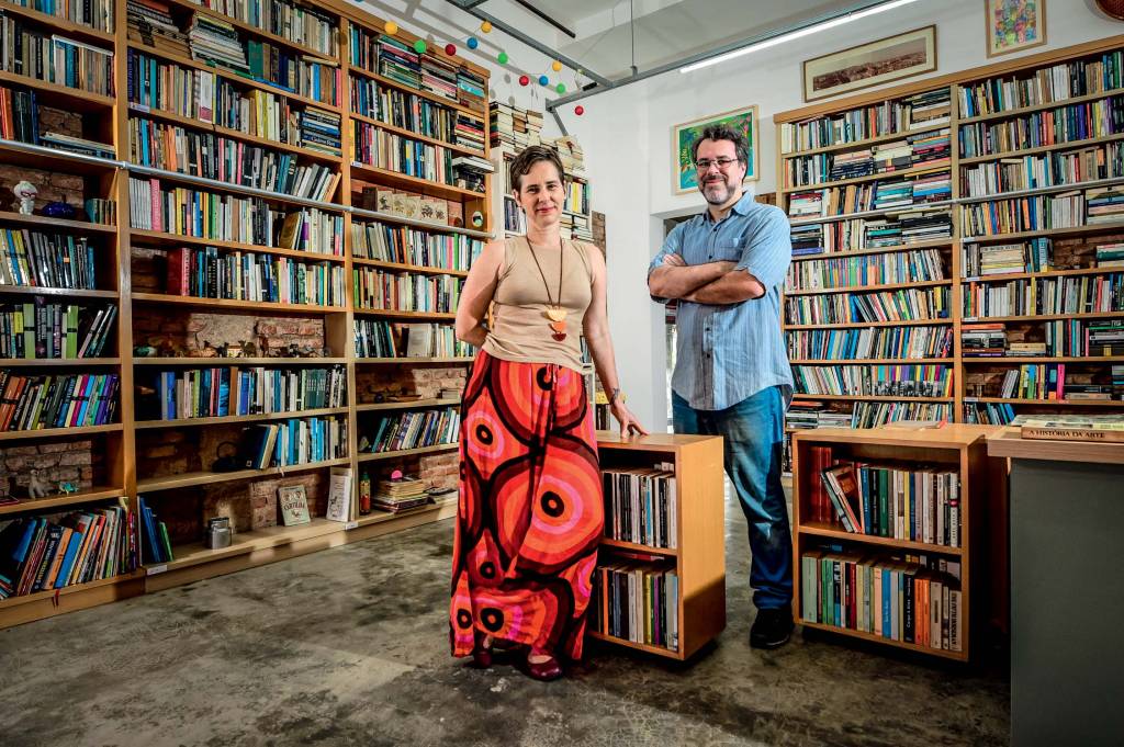 Imagem mostra sala com estantes de livros e uma mulher e um homem, ao centro, sorrindo