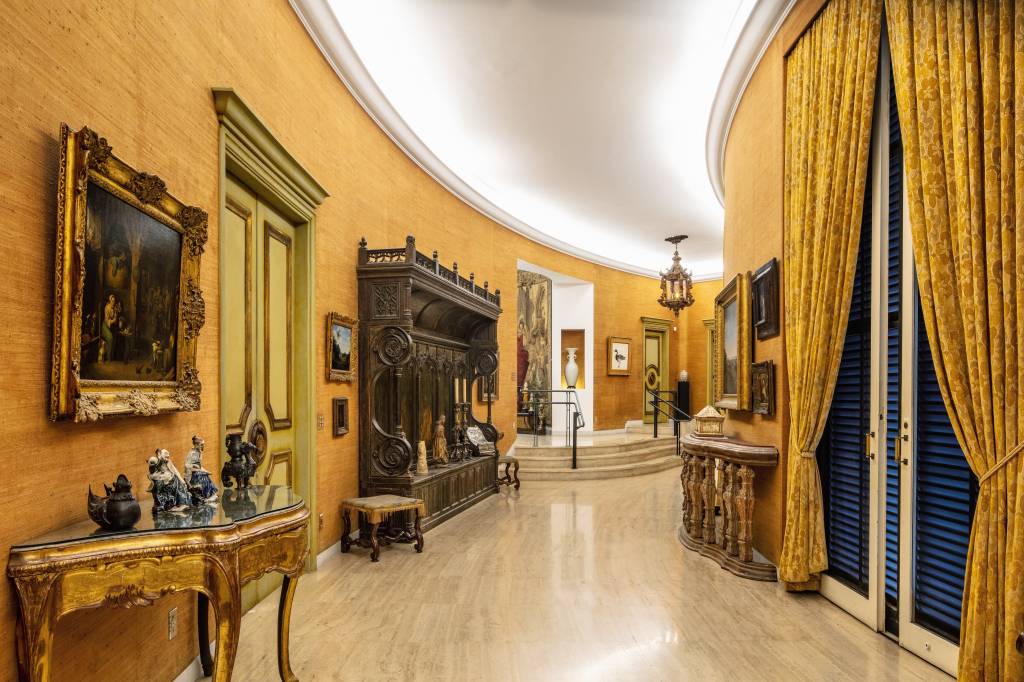 Corredor da Casa Museu Ema Klabin exibe móveis antigos e parede amarela.