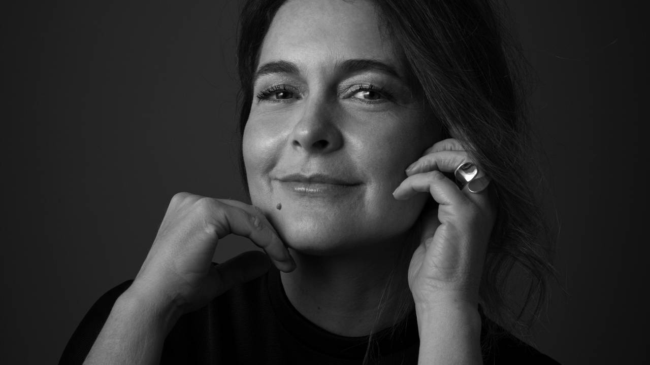 Claudia Abreu posa com as mãos no rosto, em um leve sorriso, cabelo preso. A foto está em preto e branco.