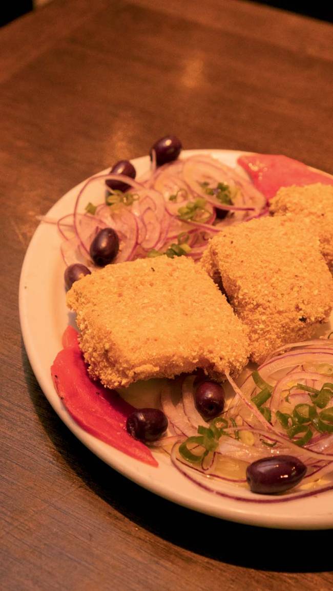 Prato de porcelana branco contendo pedaços de bacalhau empanados, saladinha de cebola-roxa, azeitona preta e pimentão vermelho