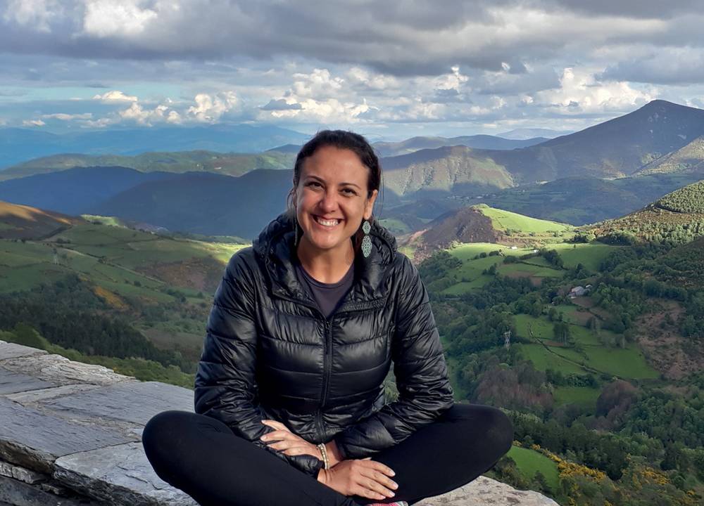 Foto mostra Gilsimara Caresia sentada de pernas cruzadas, com jaqueta bomber, em mureta, com paisagem de colinas ao fundo