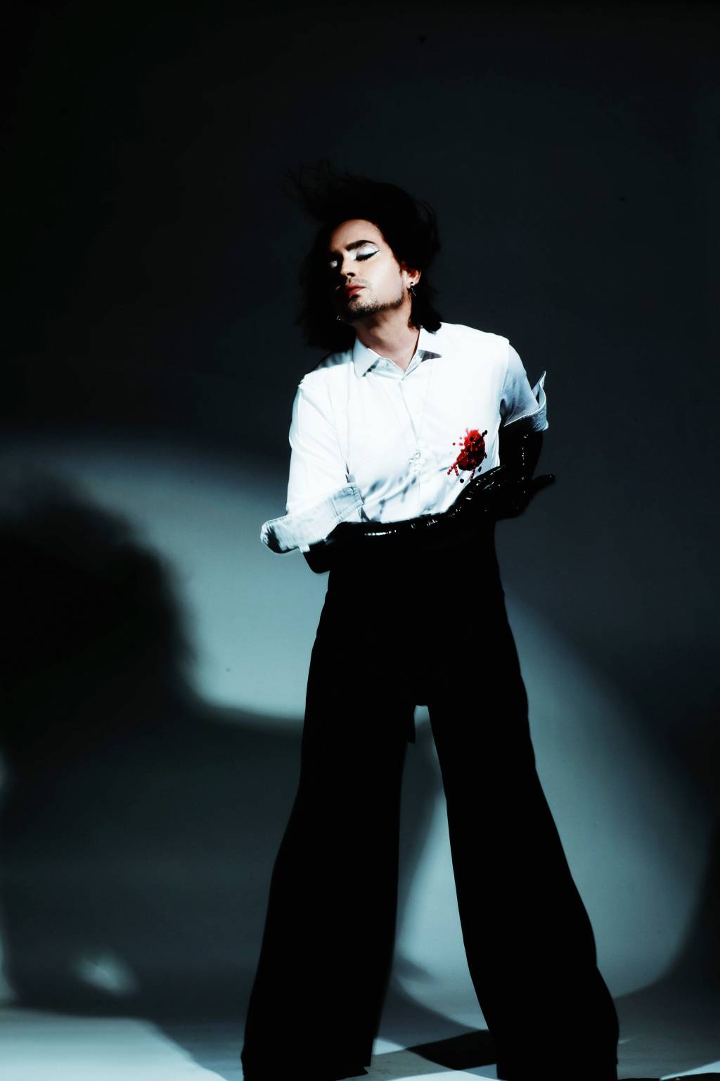 Johnny Hooker Imagem mostra homem de camisa branca e calça preta, de perfil, em pé. Na altura do coração, uma marca de sangue