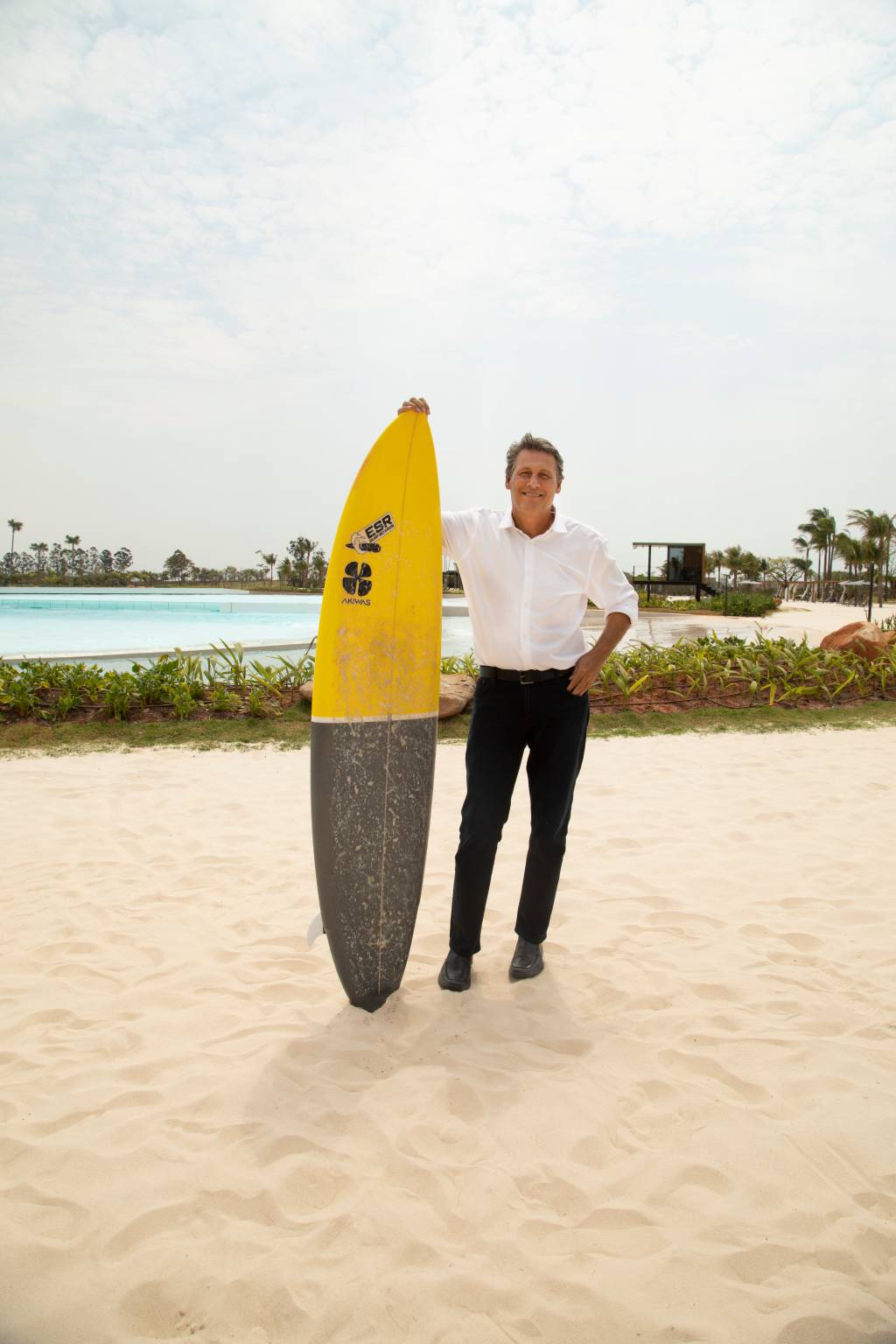 Oscar Segall (Henrique Padilha-Divulgação) (3) Homem branco grisalho veste calça preta, camisa branca e segura prancha amarela e cinza em areia com piscina ao fundo.