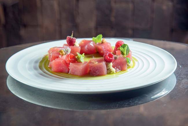 Ella Fitz crudo de atum e framboesa pistache, hortelã e stracciatella Prato de porcelana branca sobre superfície espelhada contendo cubos de atum e outros ingredientes como framboesa pistache e hortelã