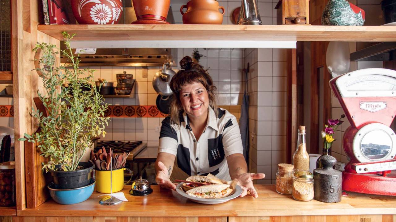 Chef do Aconchegante Bar Katia Lyra em ambiente interno do bar, sobre balcão de madeira e tendo ao fundo parede de azulejos e acima prateleira com potes de cerâmica