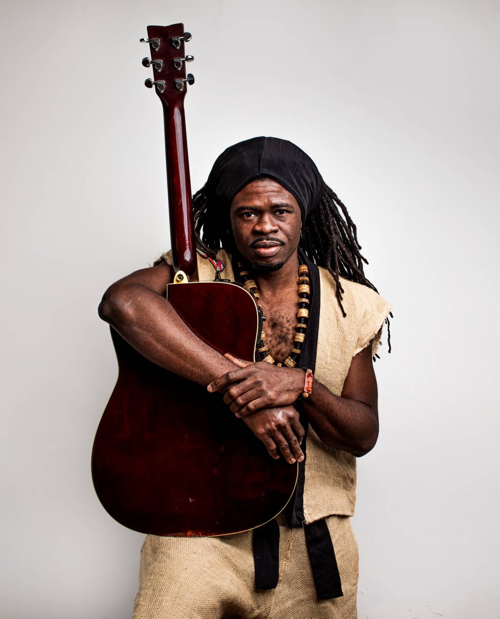 Yannick Delass (José de Holanda Estúdios-Divulgação) Cantor e compositor Yannick Delass posa abraçado com violão de pé. Tem dreads, pele negra e veste um colete cor de palha.