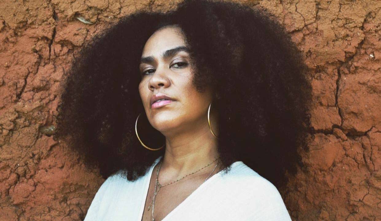 Imagem mostra mulher de roupa branca e brincos largos, encostada em parede de pedra