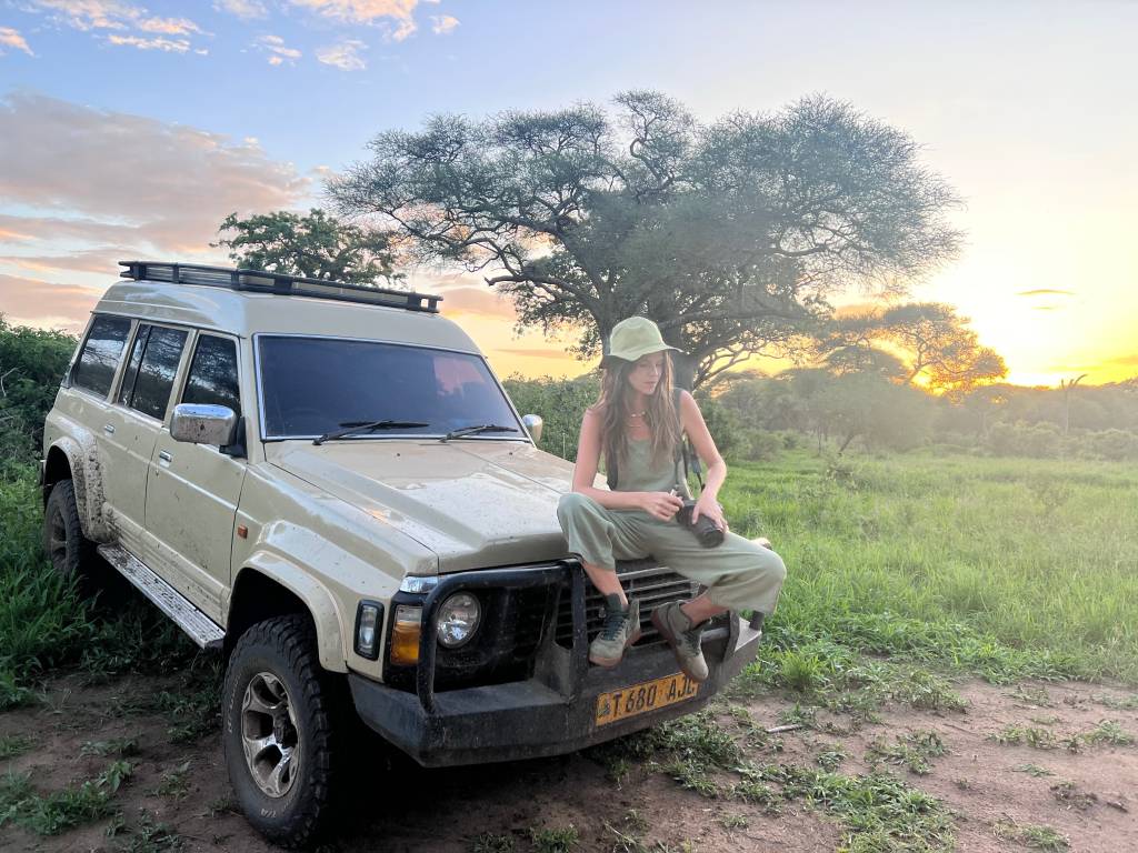 Moça branca com boné posa sentada em carro 4x4 em meio a gramado e árvore ao fundo.