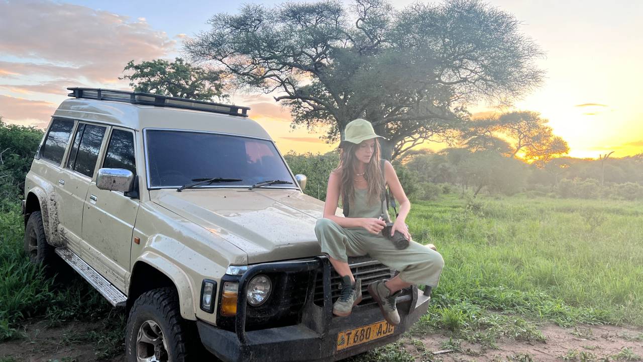 Moça branca com boné posa sentada em carro 4x4 em meio a gramado e árvore ao fundo.