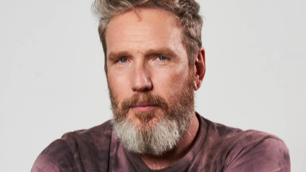 Homem branco de barba e cabelo grisalhos encara a câmera sério com os braços levemente cruzados. Veste camiseta com tom manchado na cor roxa.